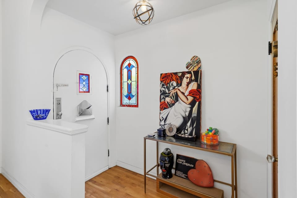 The entry foyer: stained glass arched windows, a bold art deco portrait, brass console table, and a sculptural globe pendant — the first moment that signals this home is genuinely different.