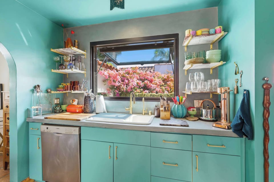 Kitchen sink wall with teal cabinetry, brass hardware, Bosch dishwasher, and a wide garden window perfectly framing a blooming bougainvillea — nature's own artwork while you do the dishes.