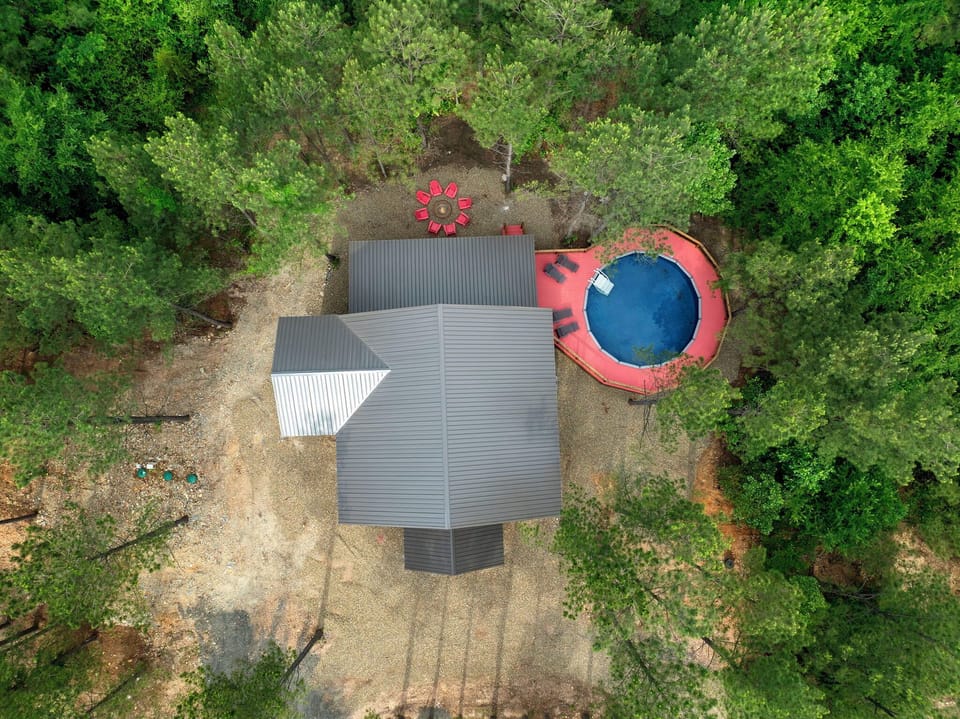 Aerial view of a charming vacation retreat nestled among lush trees, featuring a private pool with red seating area and spacious grounds perfect for outdoor relaxation.