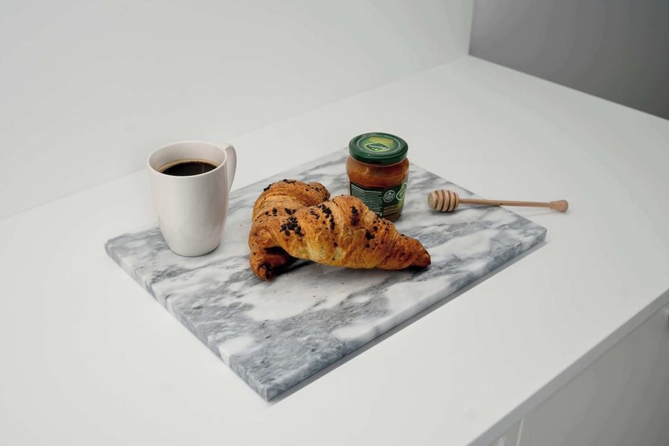 A close-up view of breakfast items arranged neatly on a marble serving board. A mug of coffee, a fresh croissant, and a small jar of jam with a spoon are featured. The bright lighting enhances the fresh and appetizing appearance of the items.