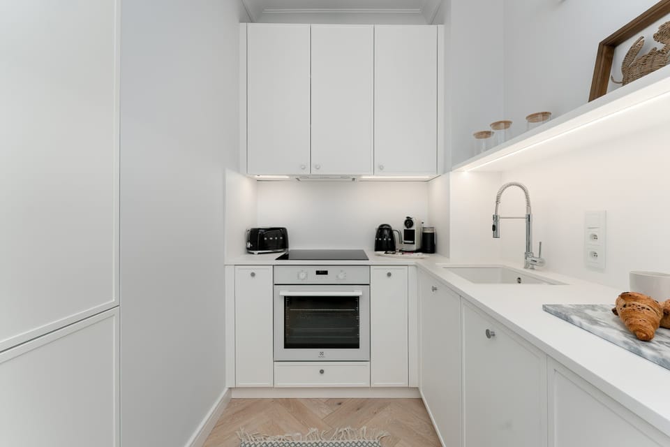 A close-up of a portion of the modern white kitchen shows the stovetop, oven, and counter space. The sleek, handles-free cabinets and a warm wooden backsplash element are prominent features. 