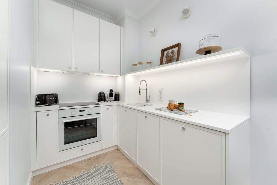 A clean, modern white kitchen is depicted from a corner perspective, showing upper and lower cabinetry. The space includes a built-in oven, stovetop, and an undermount sink. Decorative items like a potted plant and bottles are placed on the counter.