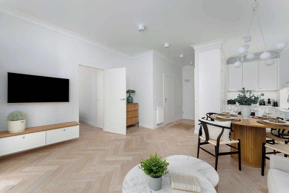 This shot provides a broad perspective of the entire apartment, showing the living area, dining set, and kitchen from across the room. The wooden flooring and grey-toned walls tie the spaces together.