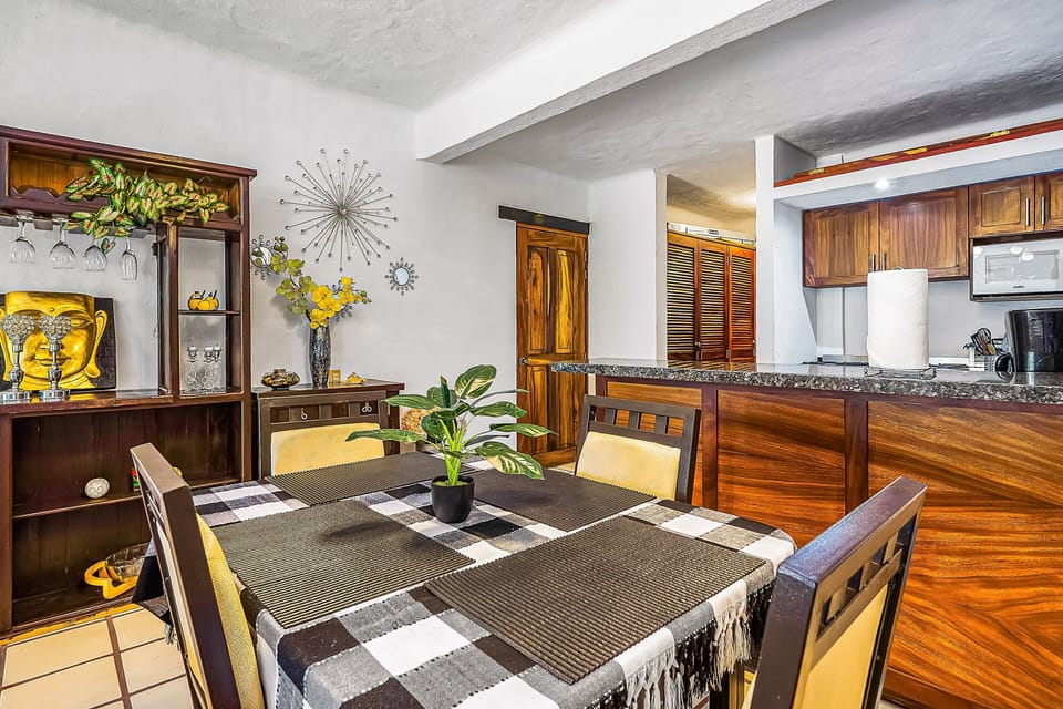 Cozy dining setup featuring a checkered tablecloth and padded chairs, accented by a decorative wooden hutch and a sleek granite bar counter that opens up directly into the kitchen.