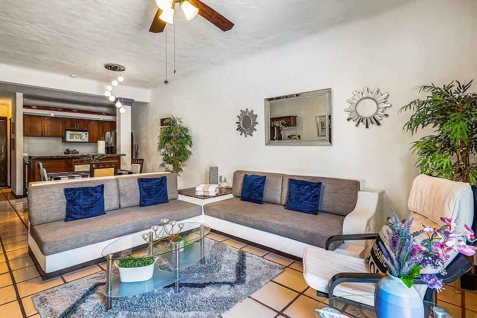 Open-concept living room with a gray sectional, glass coffee table, and stylish mirrors, flowing smoothly into a modern dining area and fully equipped kitchen just beyond.