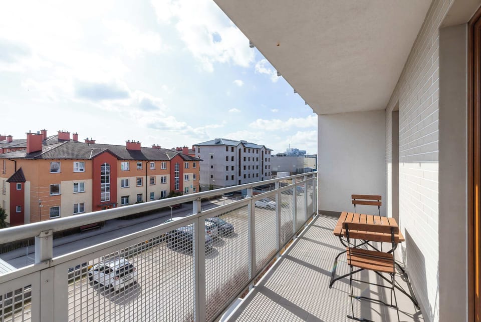 The balcony features a glass railing and offers a wide view of the surrounding residential buildings and parking area. A small wooden table and chair set provides a quiet spot for outdoor relaxation.