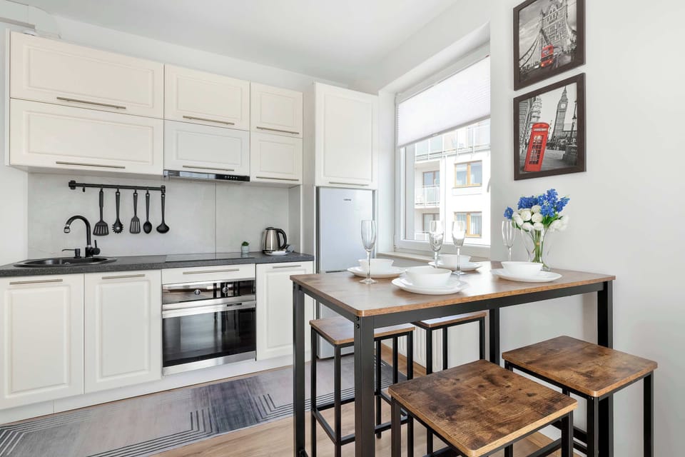A modern L-shaped kitchen is equipped with white cabinets, dark countertops, and integrated appliances like a stove and oven. A dark wooden dining table with bar stools is positioned nearby for casual meals.