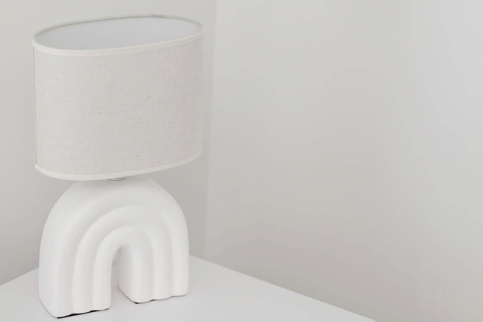 A small white lamp with a unique, textured arch-shaped base sits on a plain white table. The minimalist setting against a neutral wall highlights the lamp's modern and artistic design.
