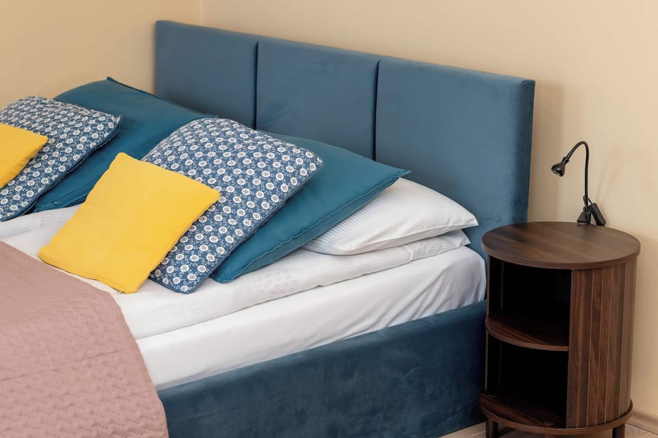 A close-up of the bedding reveals high-quality blue linens topped with vibrant yellow and patterned cushions. A modern black reading lamp stands on the dark wood bedside table for evening convenience.