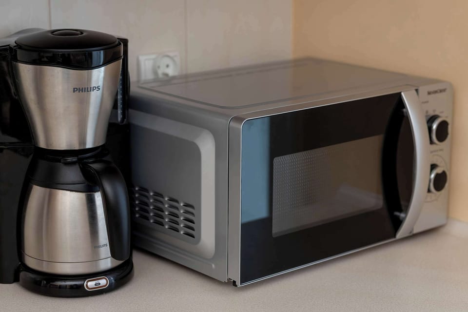A compact kitchenette workstation holds a sleek silver microwave and a drip coffee machine ready for use. The layout is designed for efficiency, keeping essential appliances within easy reach.