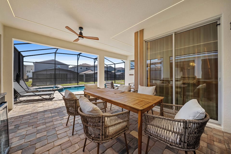 Enjoy the inviting outdoor dining area with a rustic wooden table and stylish woven chairs. Large windows let in natural light, while a ceiling fan provides a refreshing breeze as you savor meals by the pool.