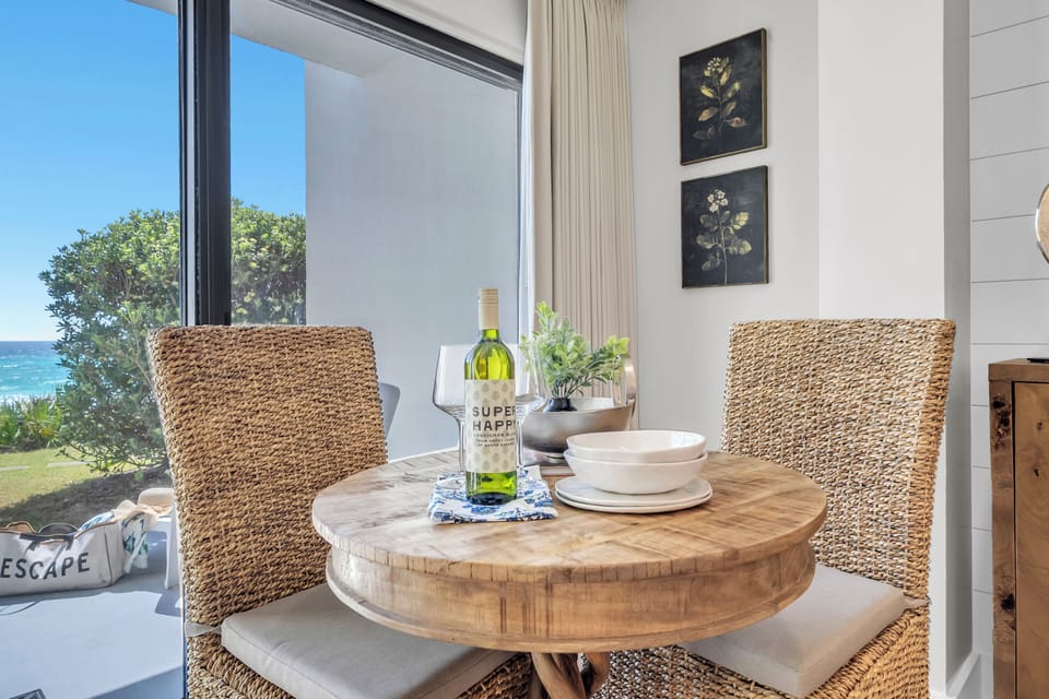 Your intimate dining spot showcases stunning ocean views through floor-to-ceiling windows, where wicker chairs embrace a rustic wooden table perfect for romantic meals.