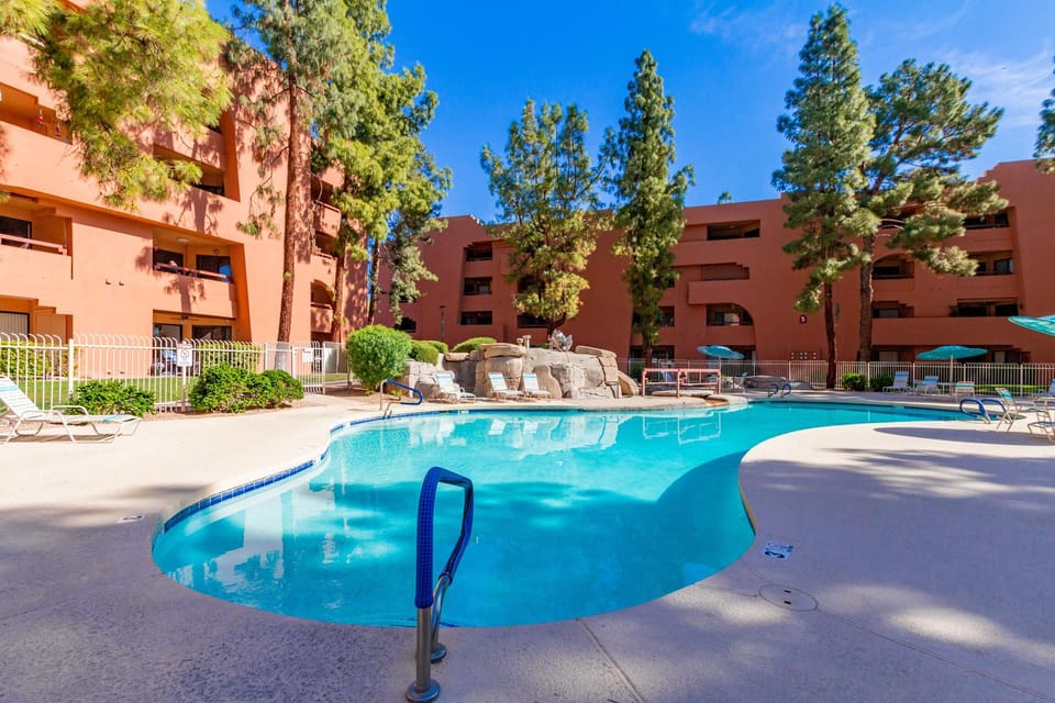 Curved outdoor pool with rock waterfall, surrounded by lounge chairs and terracotta-colored buildings.
