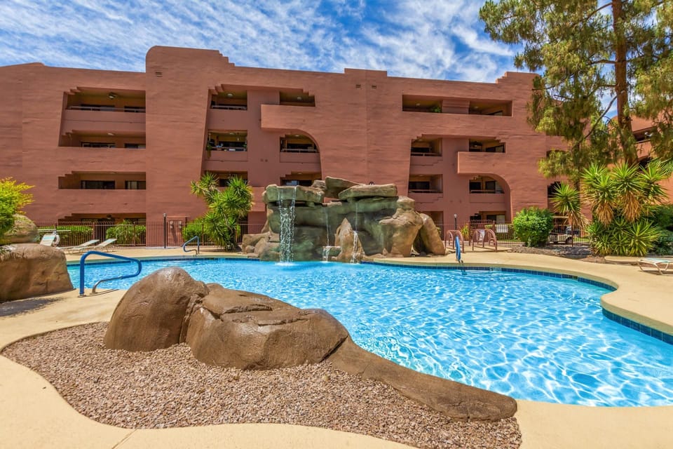 Curved pool edged with tan concrete and natural rock waterfall sits in front of terracotta stucco condo building.