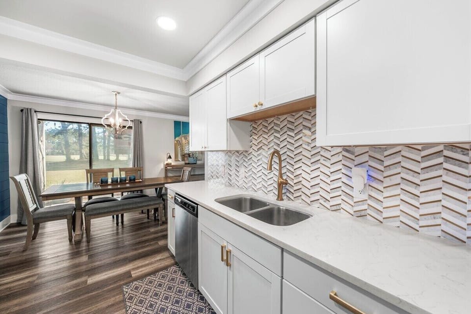 Bright kitchen with white shaker cabinets, quartz counters, gold chevron backsplash, and double sink — dining table for six steps away