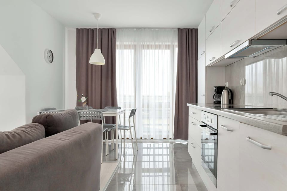 A bright dining corner is situated next to a window, featuring a white table with transparent chairs and a nearby kitchenette.