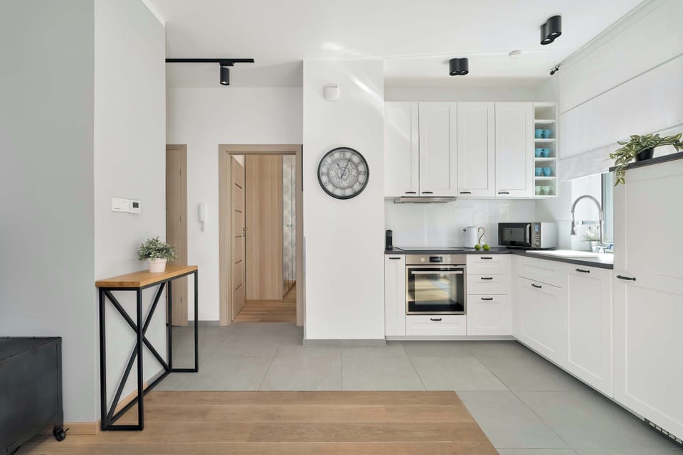 A minimalist white kitchenette is equipped with modern handleless cabinetry, a built-in oven, and sleek black track lighting on the ceiling.