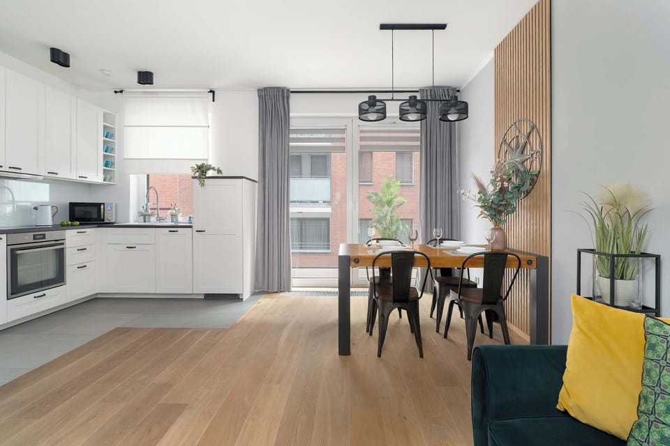A kitchenette and dining perspective captures the bright, airy feel of the room, looking from the white cabinets toward the wooden table and chairs.