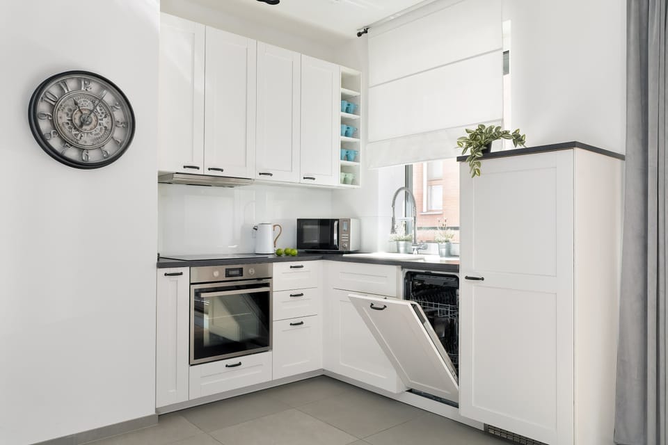 A functional kitchenette corner features an L-shaped arrangement with white cabinetry, an integrated dishwasher, and a large wall clock.