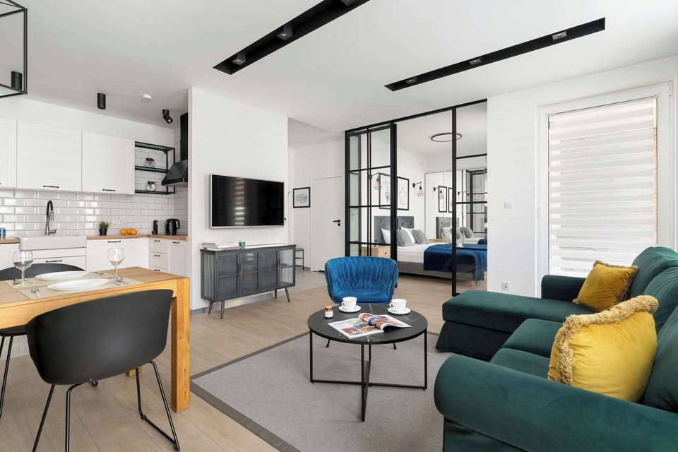 A wide view of the open-plan living area reveals a comfortable green sofa and blue velvet chairs, seamlessly connected to a modern kitchen with white cabinetry. Large windows let in plenty of natural light.