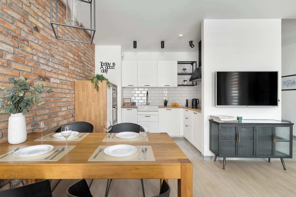 The modern kitchen with its clean white cabinets and dark countertops is partially visible from the living area. The exposed brick wall transitions to a white wall featuring a mounted television and a sleek storage unit.