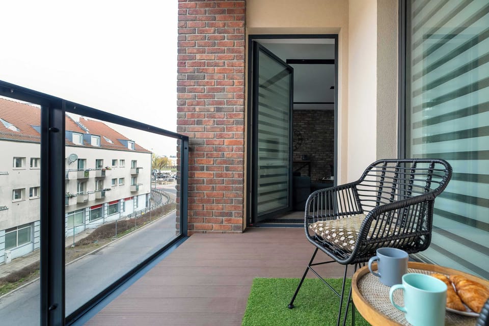 A modern balcony provides an outdoor retreat with a stylish black woven chair and a small side table holding two mugs. A wooden deck and glass railing overlook other buildings, creating a pleasant cityscape view.