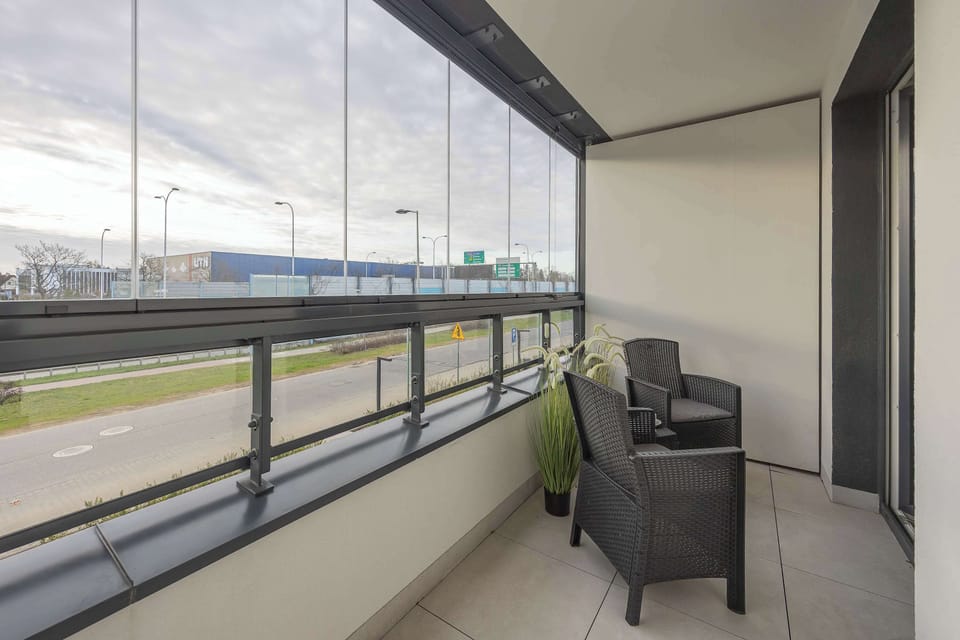 A balcony with outdoor seating, including chairs and a small table. Glass railings provide an unobstructed view of the surroundings. The space is ideal for relaxing outside.