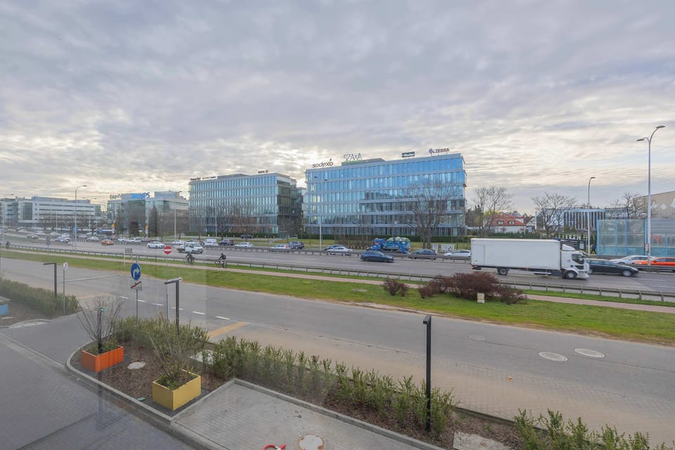 A view from a balcony overlooking a street and modern buildings. The scene includes greenery and a cloudy sky. The perspective highlights the surrounding neighborhood.