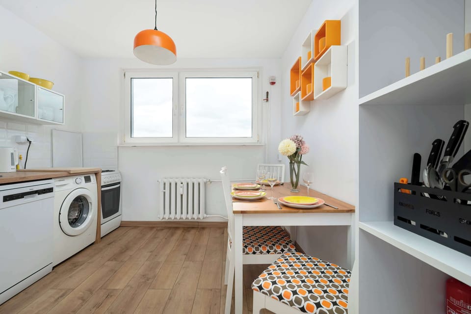 A bright kitchen corner with a small dining table placed by the window. Orange details in the decor and lighting add a cheerful touch to the space. Natural light enhances the clean, functional design.