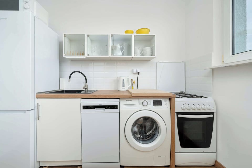 A compact kitchen area with white cabinets, a countertop, and a washing machine integrated below. Open shelves above hold simple kitchen items, keeping everything within easy reach. The space feels practical and neatly arranged.
