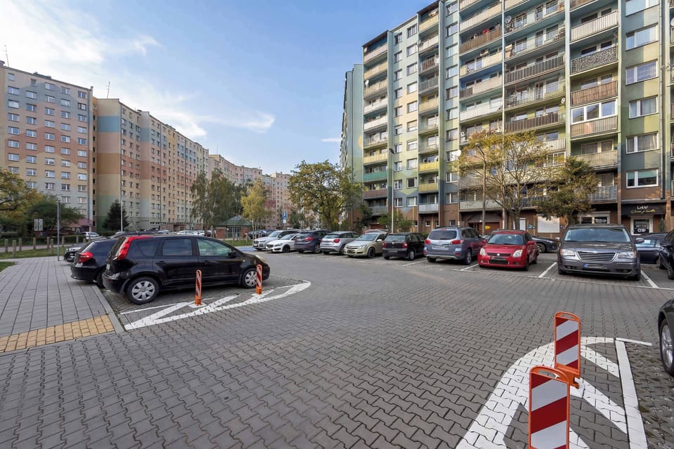 A street-level view of the building with parked cars and a paved walkway. Surrounding apartment blocks suggest a residential neighborhood. The area looks orderly and easily accessible.