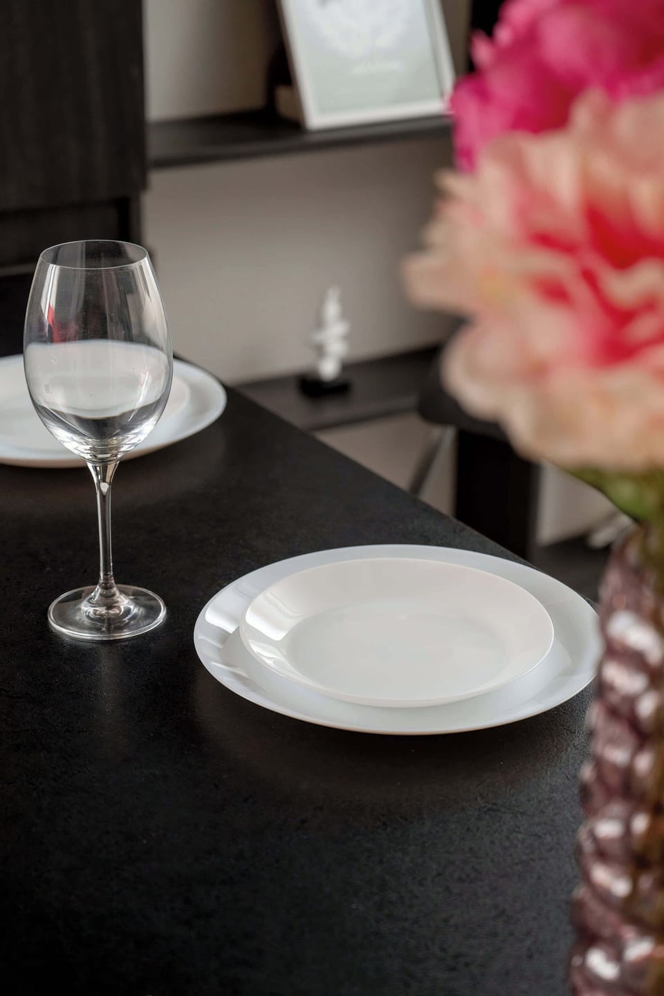 A table setting detail features a white ceramic plate and a wine glass, set against the backdrop of vibrant pink flowers.