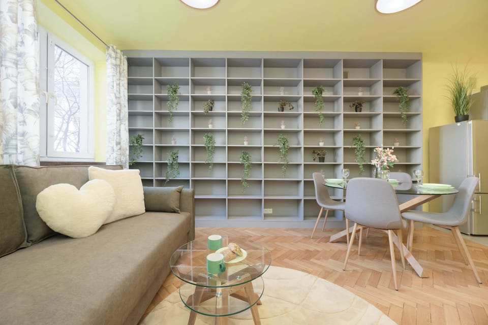 A spacious living area combining a comfortable green sofa, a coffee table with a plant, and a large dining table with modern chairs, set against yellow walls and bookshelves.
