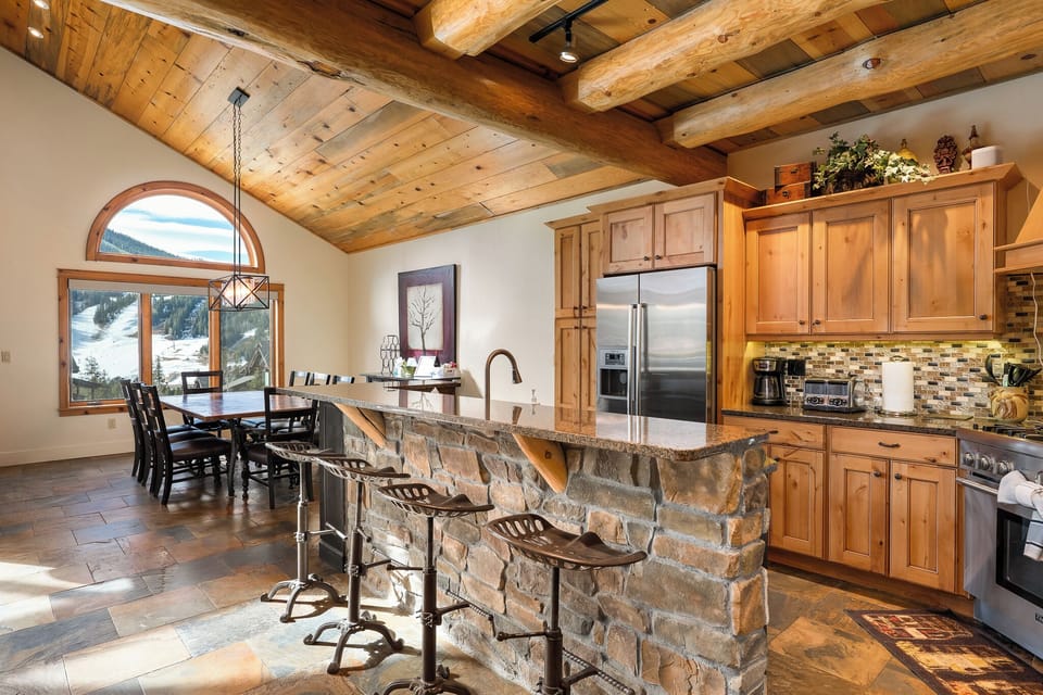 Gather in your stunning mountain kitchen with exposed wood beams, stone island, and panoramic ski slope views through arched windows.