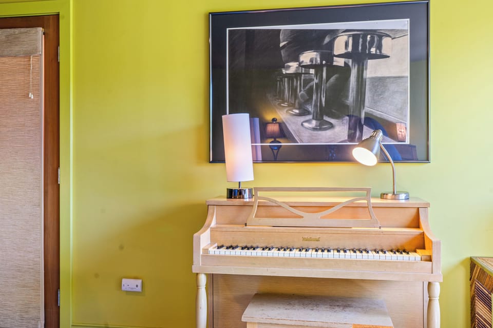 Artistic detail in "The Crag's" Base Camp bedroom. A blonde wood piano, warm directional lamps, and striking framed art create a unique, creative corner inside this ground-floor retreat near Acadia.