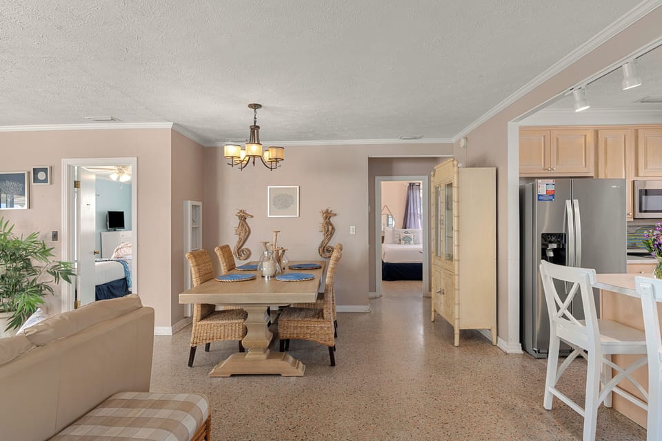 Bright coastal dining and kitchen area with woven seating, beach‑inspired décor, and an easy flow into two cozy bedrooms.