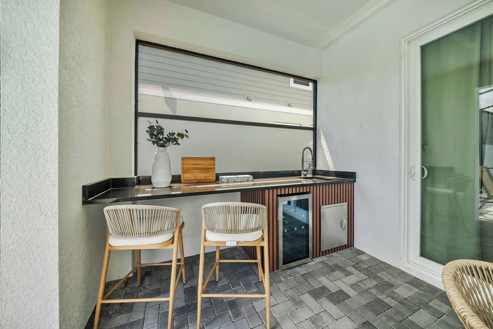 Experience a chic outdoor kitchen with sleek black countertops and modern wood cabinetry. This bright space features high-backed stools and a large window, ideal for savoring delicious meals while enjoying the fresh air.