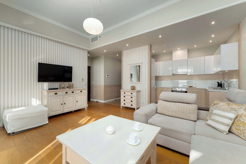 A bright open-plan living room connected to a modern kitchenette featuring a comfortable sofa, a white coffee table, and a TV area. The wooden flooring adds warmth, while recessed ceiling lights provide even illumination. 