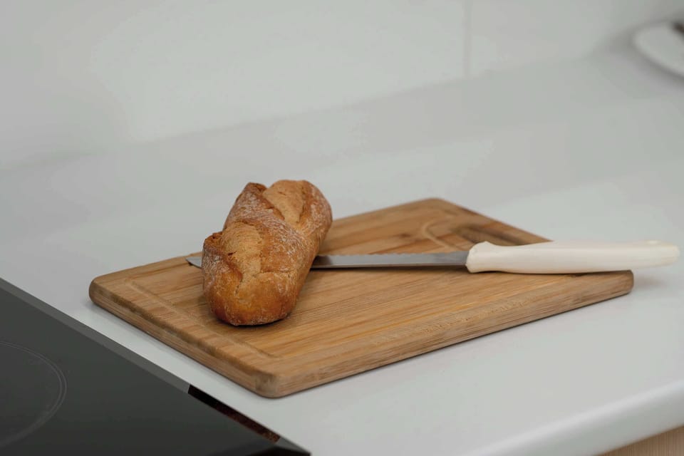 A kitchen countertop with a wooden cutting board and a fresh bread placed on it. The surface is clean and minimal, emphasizing a simple and aesthetic presentation. The warm tones add a touch of comfort.