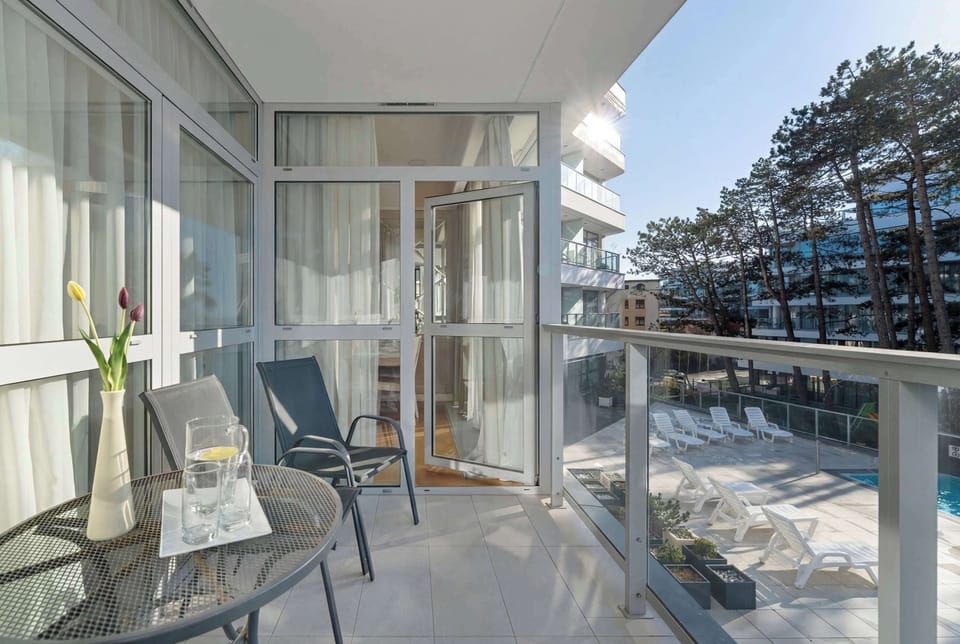 A wider view of the outdoor pool area with multiple sun loungers arranged neatly around it. Tall trees and modern buildings frame the space, creating a resort-style environment. The setting feels open, sunny, and inviting.
