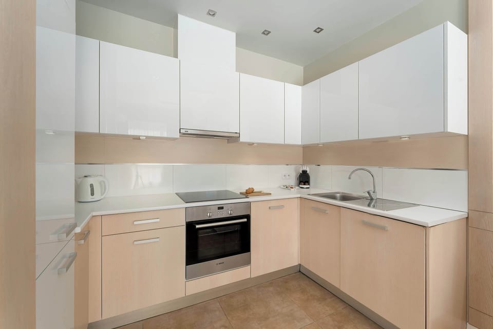 A clean and modern kitchenette with white upper cabinets and light wood lower cabinets arranged in an L-shape. Built-in appliances, including an oven and stovetop, are seamlessly integrated into the design.