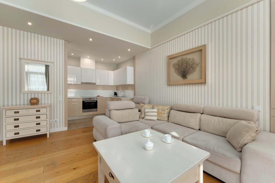 A spacious living room with a large L-shaped sofa arranged around a white coffee table, creating a comfortable area for relaxation. Vertical striped wallpaper and a framed artwork add a subtle decorative touch.