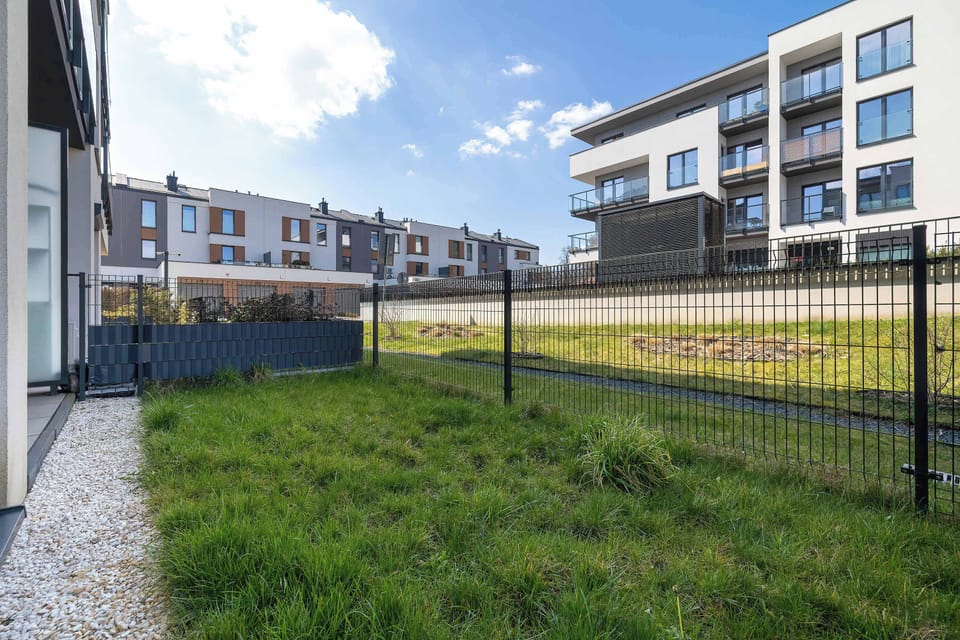 A fenced-in green garden area provides a private outdoor space for the ground-floor apartment. The well-maintained lawn is bordered by the modern facade of the residential building. 