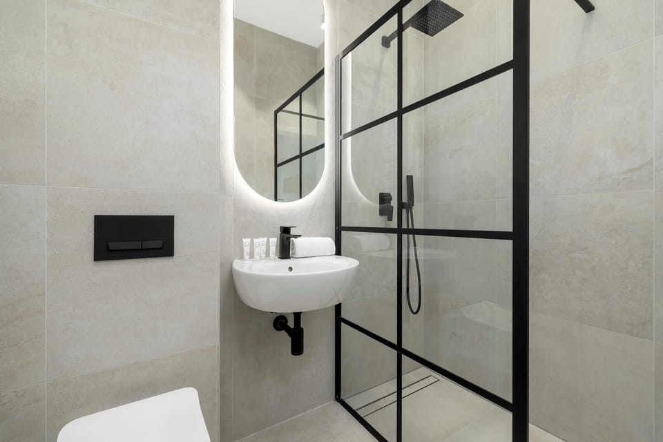 A clean bathroom with light gray tiled walls, a sink with a mirror, and a black-framed glass shower enclosure.