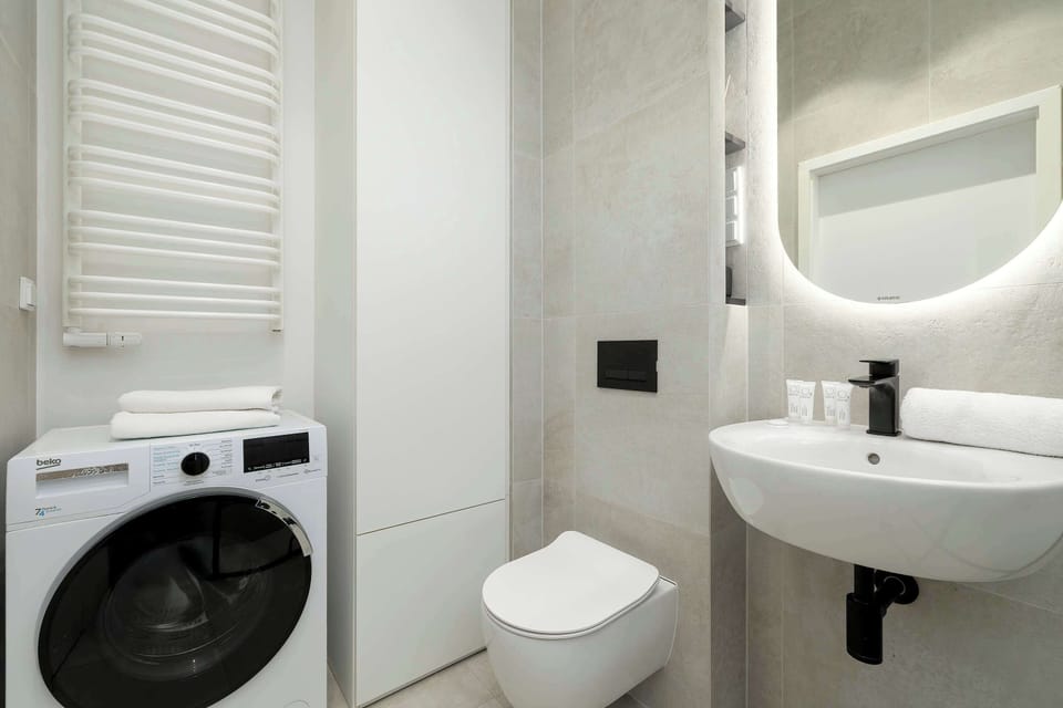 The bathroom features a top-loading washing machine placed next to the vanity for maximum utility. A backlit circular mirror provides soft illumination above the sink area.