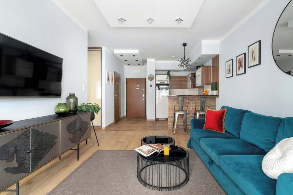 A vibrant teal sofa sits opposite a large television in this modern open-plan living and kitchenette area with  white cabinetry and dark countertops, separated by a brick-patterned breakfast bar with two metal stools.