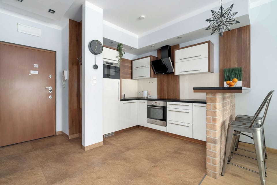 Dark-patterned floor tiles unify the L-shaped modern kitchenette and adjacent entryway. White cabinets and darker appliances define the cooking space, which is anchored by a kitchen island with a dark top and two metal bar stools. 
