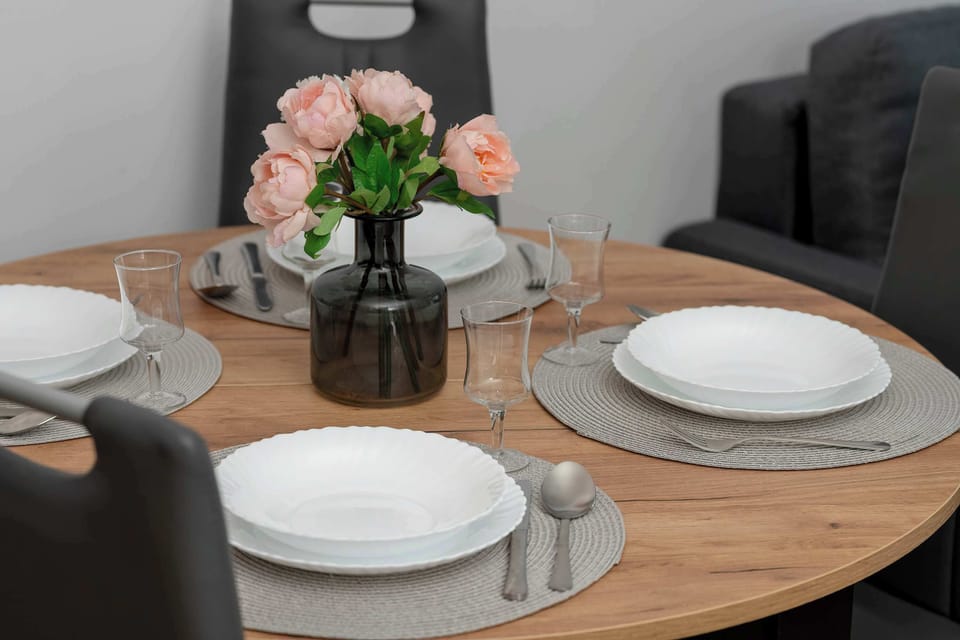 A close-up of a dining table set with plates, napkins, and a vase of pink flowers as a centerpiece.
