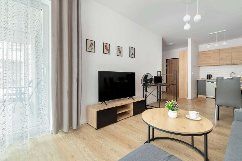 A living space with a TV, coffee table, and open layout leading into a kitchen area in the background.