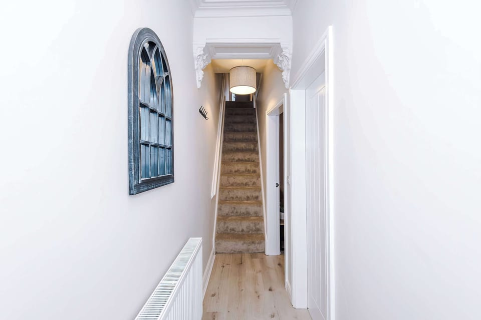 A bright, minimalist hallway leading to a carpeted staircase, with light walls, wooden floors, and a decorative arched mirror above a radiator. Clean lines and symmetry create a welcoming, modern interior.